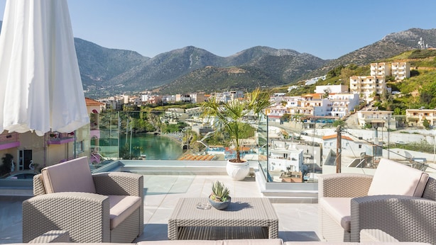 A rooftop terrace with wicker furniture, a potted plant, and a view of a swimming pool and buildings in the background. - Akrogiali Apartments (Mylopotamos)