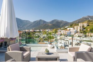 A rooftop terrace with wicker furniture, a potted plant, and a view of a swimming pool and buildings in the background. - Akrogiali Apartments (Mylopotamos)