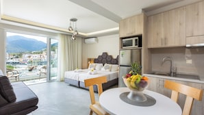A modern hotel room with a balcony, a bed with a grey headboard, a dining area with a white table, and a kitchen with wooden cabinets and a stainless steel sink. - Akrogiali Apartments (Mylopotamos)