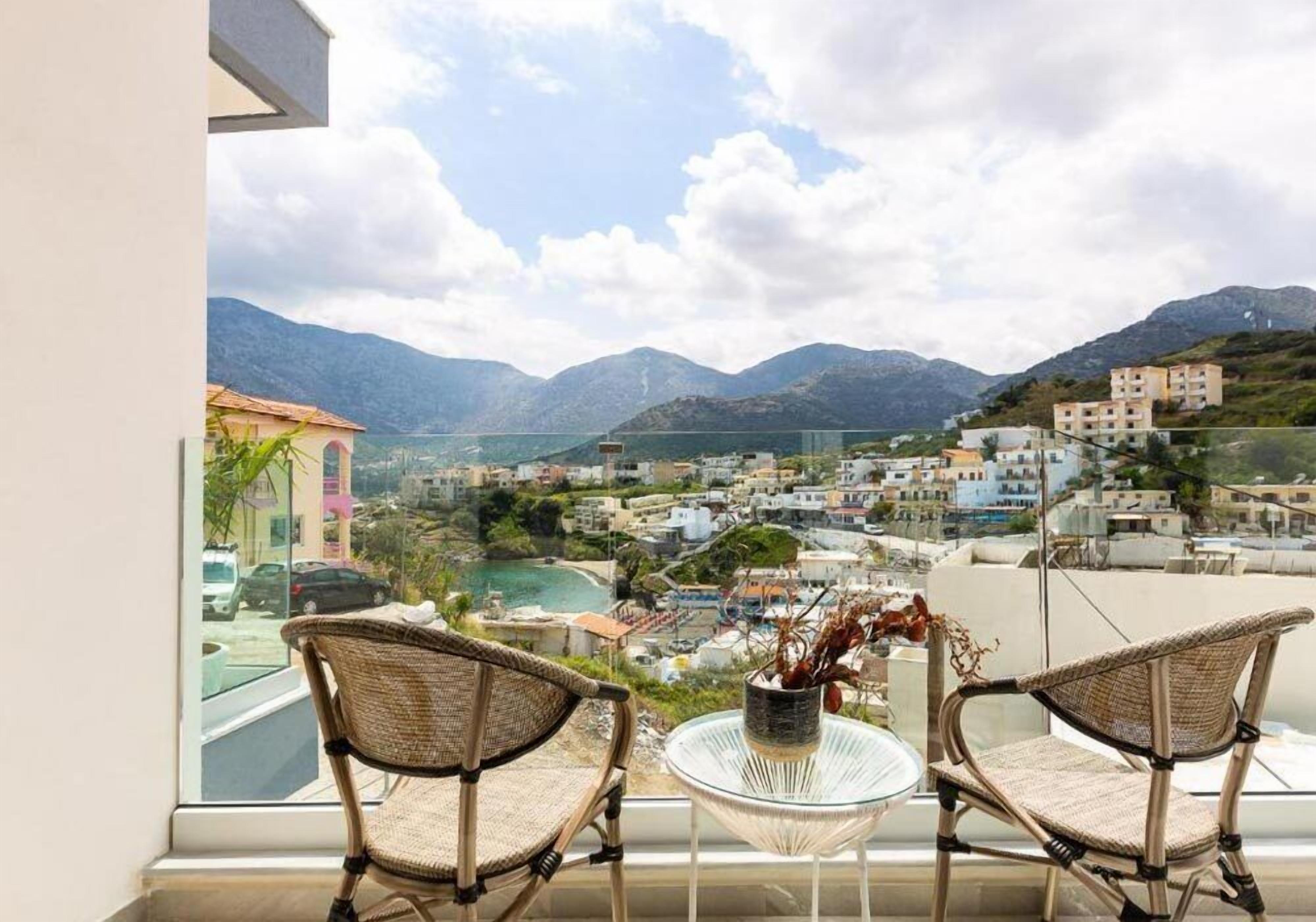 A balcony with wicker chairs and a glass table, overlooking a coastal town with mountains in the background.