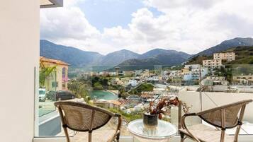 A balcony with wicker chairs and a glass table, overlooking a coastal town with mountains in the background.