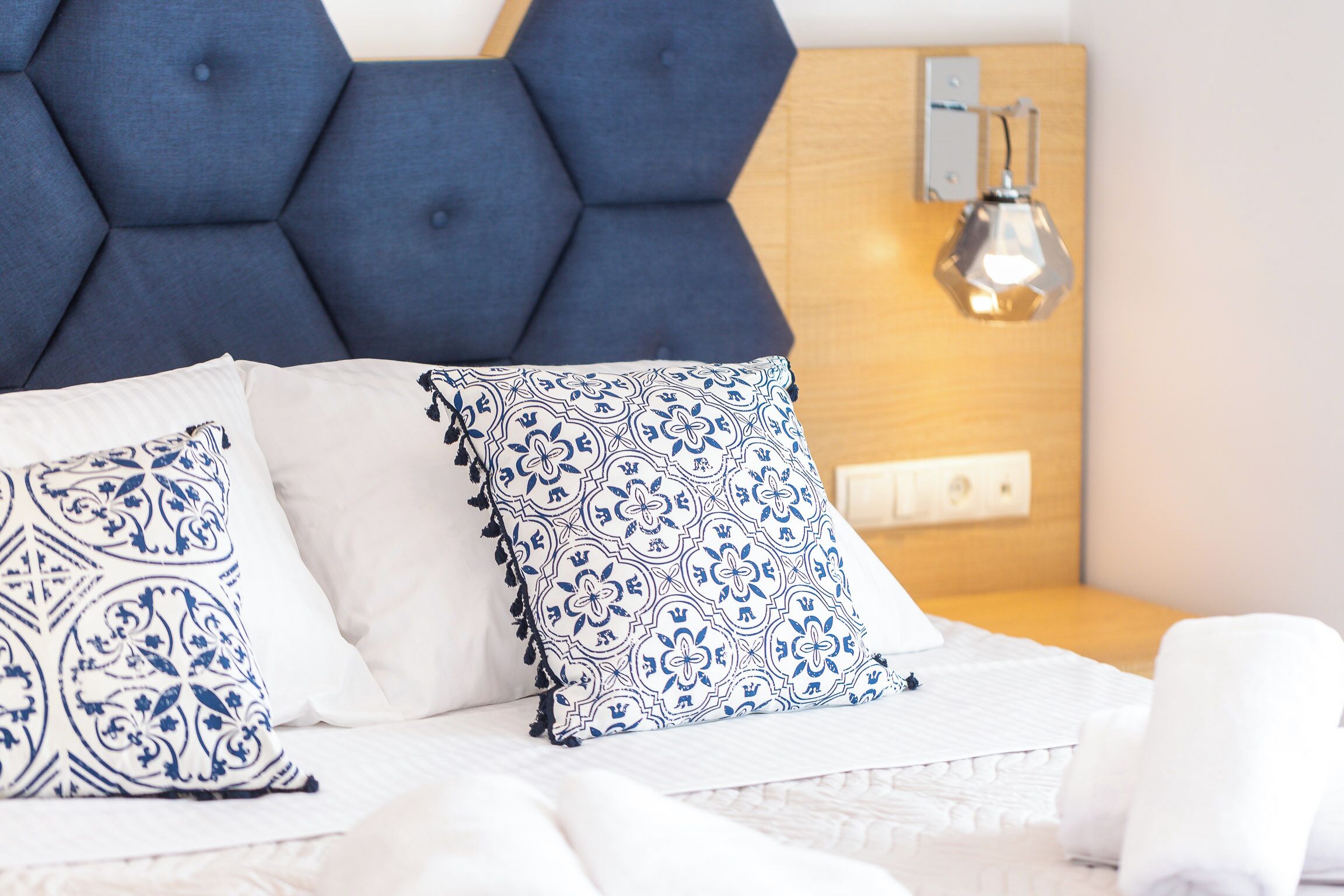 A bed with white bedding, two decorative pillows, a blue headboard, and a wall-mounted light fixture.