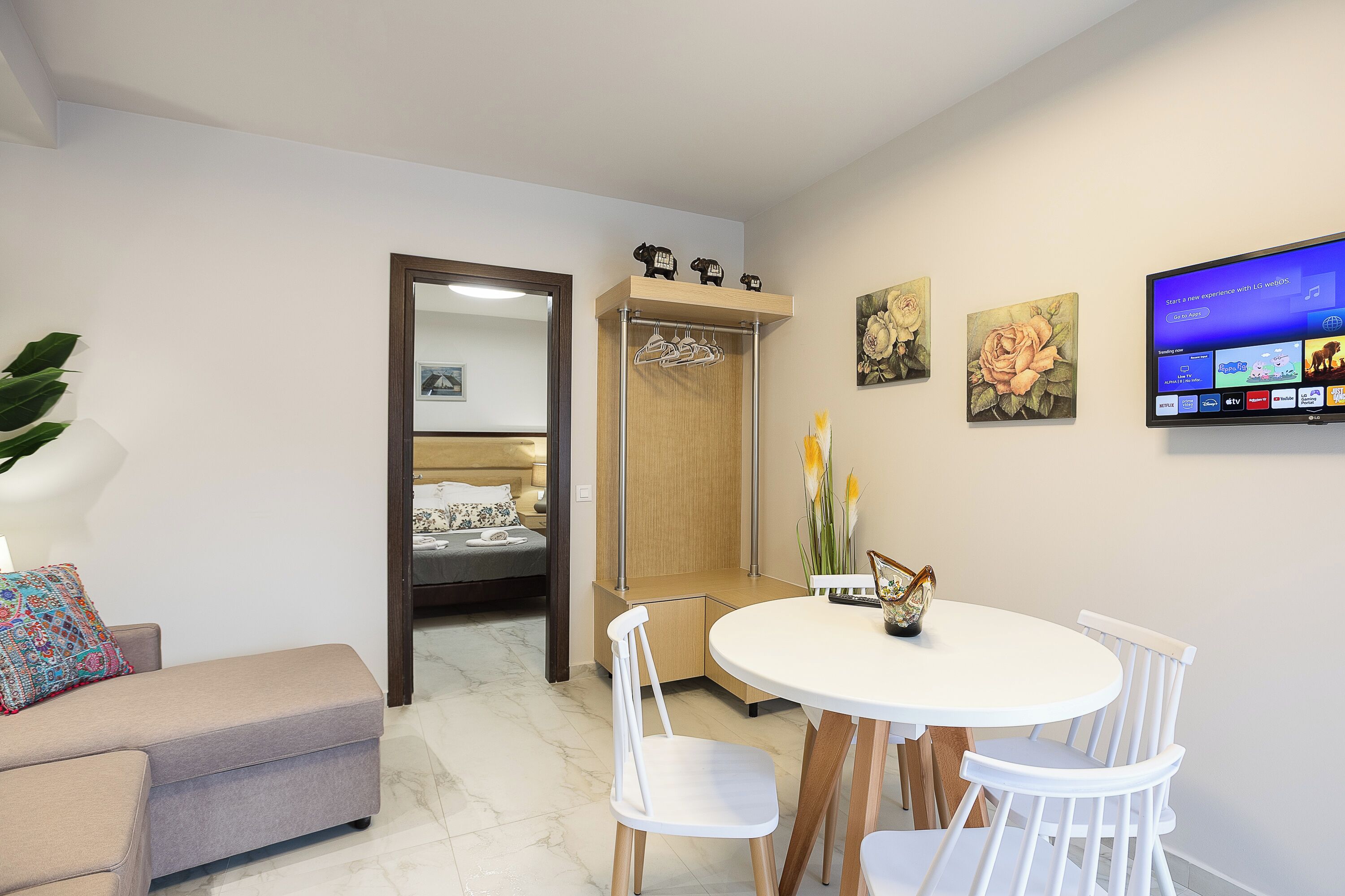 Un salon moderne comprenant un canapé, une table à manger avec des chaises, un téléviseur fixé au mur et un meuble en bois où sont suspendus des vêtements.