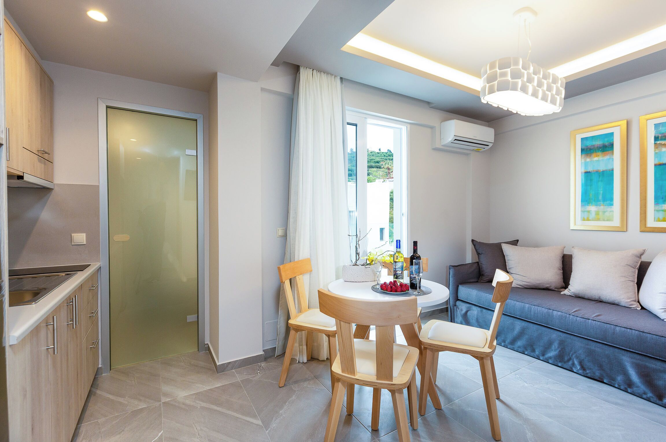 A modern kitchen with a dining area, a sofa, and a view of greenery outside.