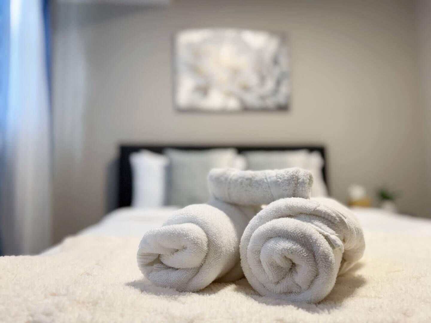 A neatly made bed with rolled white towels and a plain wall with a single piece of abstract art.