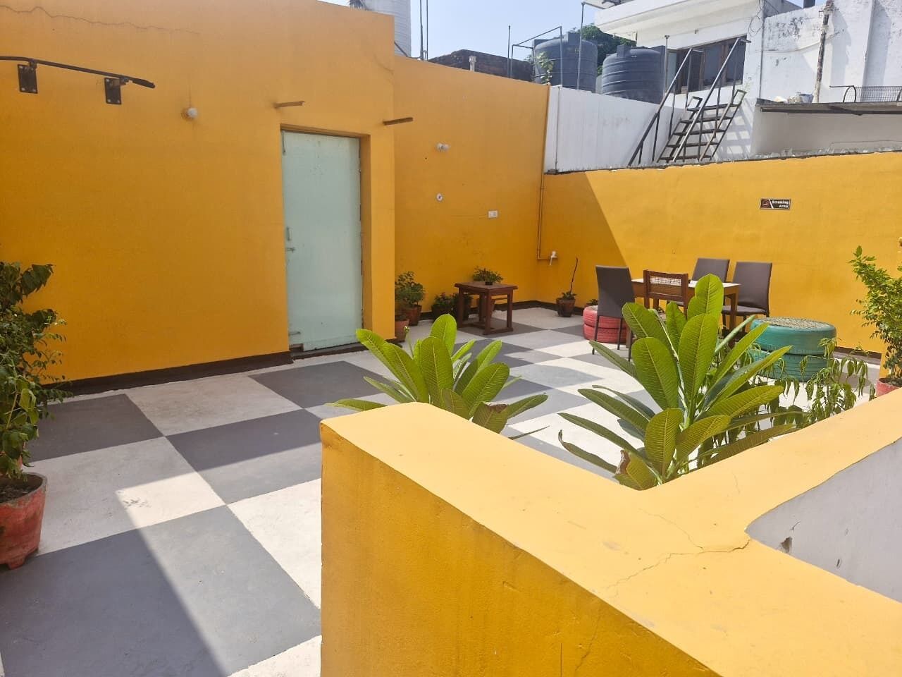 A yellow-walled outdoor area with a checkered floor, potted plants, and a table with chairs.