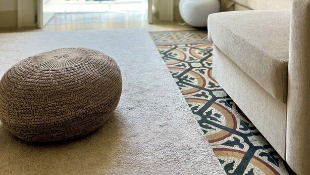 Een woonkamer met een beige bank, een rond geweven pouf en een kamerkleed met een patroon. Door een open deur is er uitzicht op gebouwen en een groene hekken.'