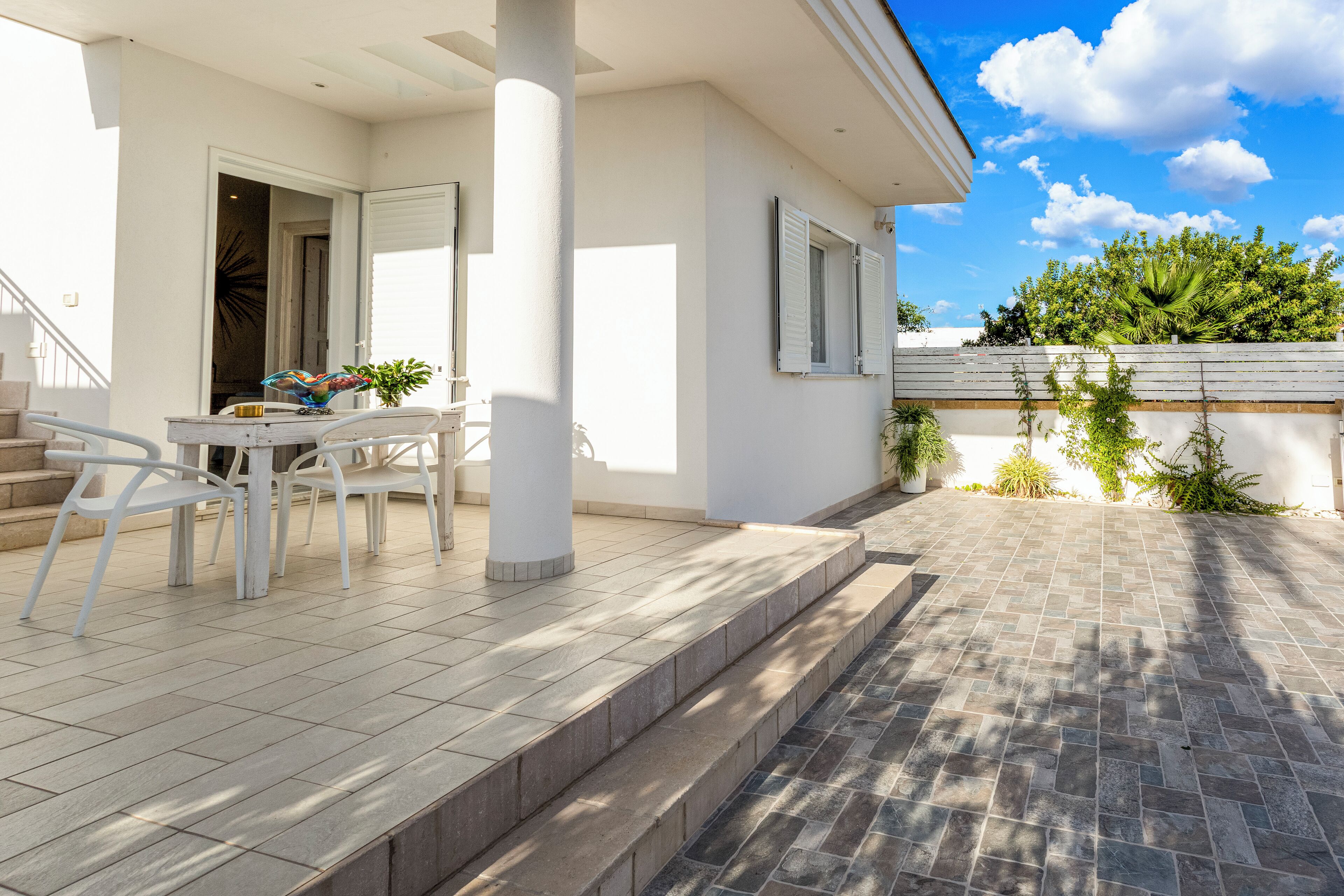 A modern house with a patio area featuring a white table and chairs, a small garden, and a view of the sky and trees.