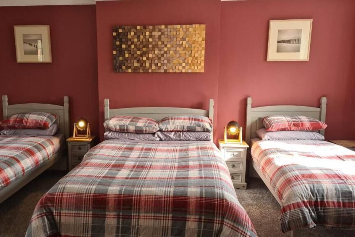 Quarto com três camas de solteiro, cada uma com colcha xadrez e almofadas a combinar. Há mesas de cabeceira com abajures, e as paredes são pintadas em um tom de vermelho quente.