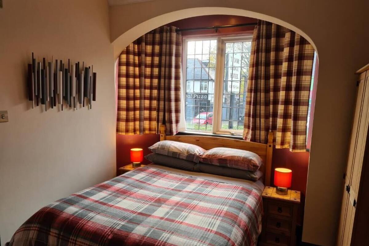Quarto com colcha xadrez, duas luminárias de cabeceira, janela com cortinas e luminária fixada na parede.