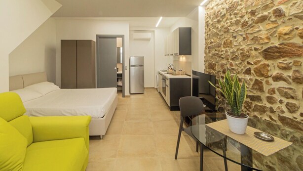 A compact hotel room with a bed, a small kitchenette, a dining area, and a stone accent wall.
