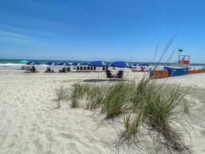 A beach with rows of blue umbrellas and beach chairs, a lifeguard tower, and the ocean in the background.