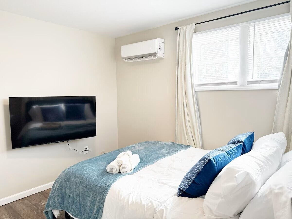 Una habitación de hotel con una cama, un televisor de pantalla plana montado en la pared y un equipo de aire acondicionado sobre la cama.