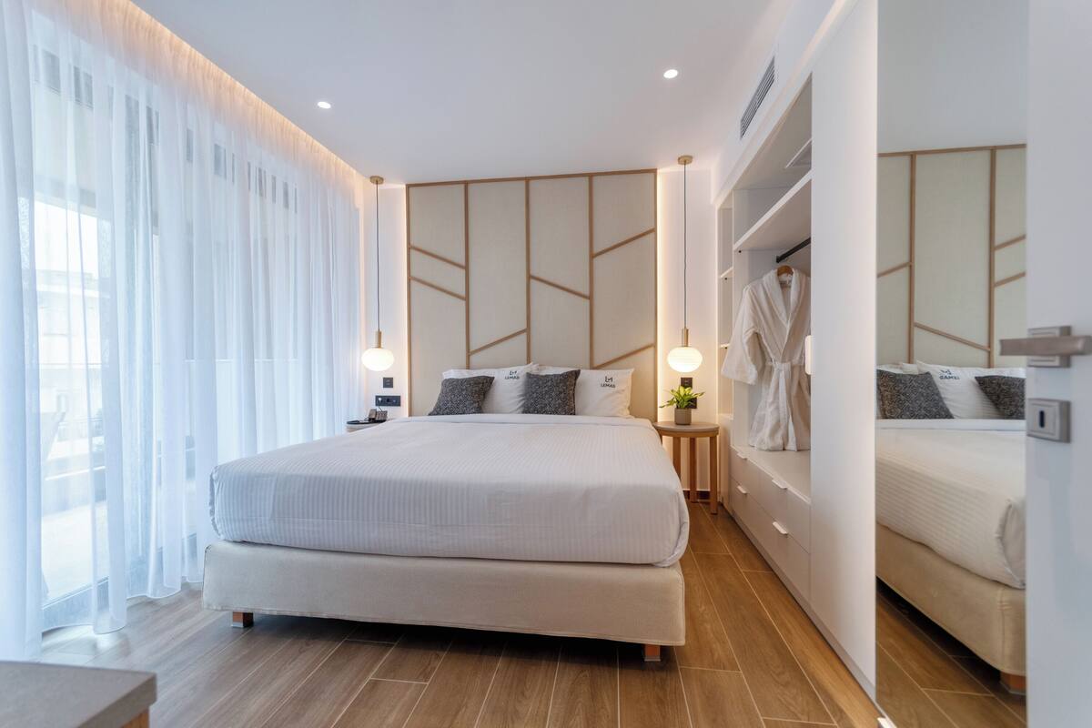 Une chambre d’hôtel moderne, dotée d’un grand lit, de tables de chevet, d’une armoire et d’une grande fenêtre avec des rideaux légers.