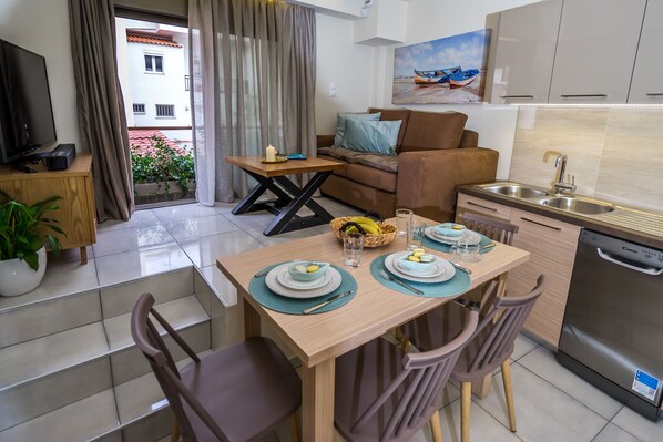 A modern kitchen with a dishwasher, a dining table set with plates and cutlery, a sofa, and a TV in the living area.