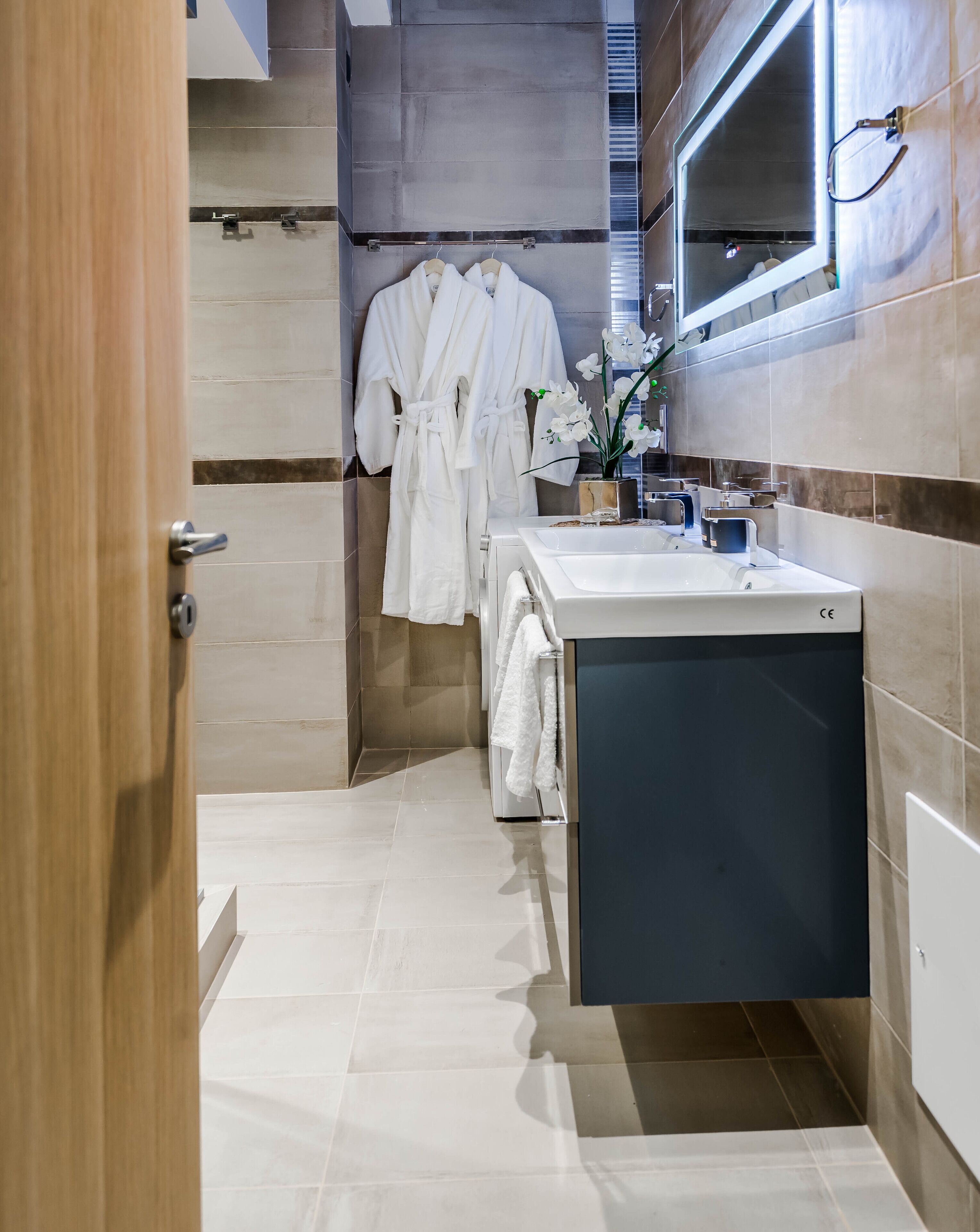 Un bany modern amb un lavabo, un mirall i dues albornoces blanques penjades a la paret.