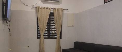 A room with a flat-screen TV mounted on the wall, a ceiling fan, a window with curtains, a dark-colored sofa, and a tiled floor.