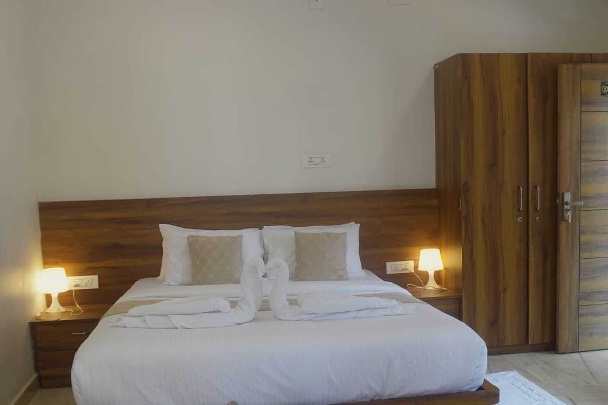 A neatly made bed with white linens and two pillows, flanked by bedside lamps, in a room with wooden flooring and a large wardrobe.