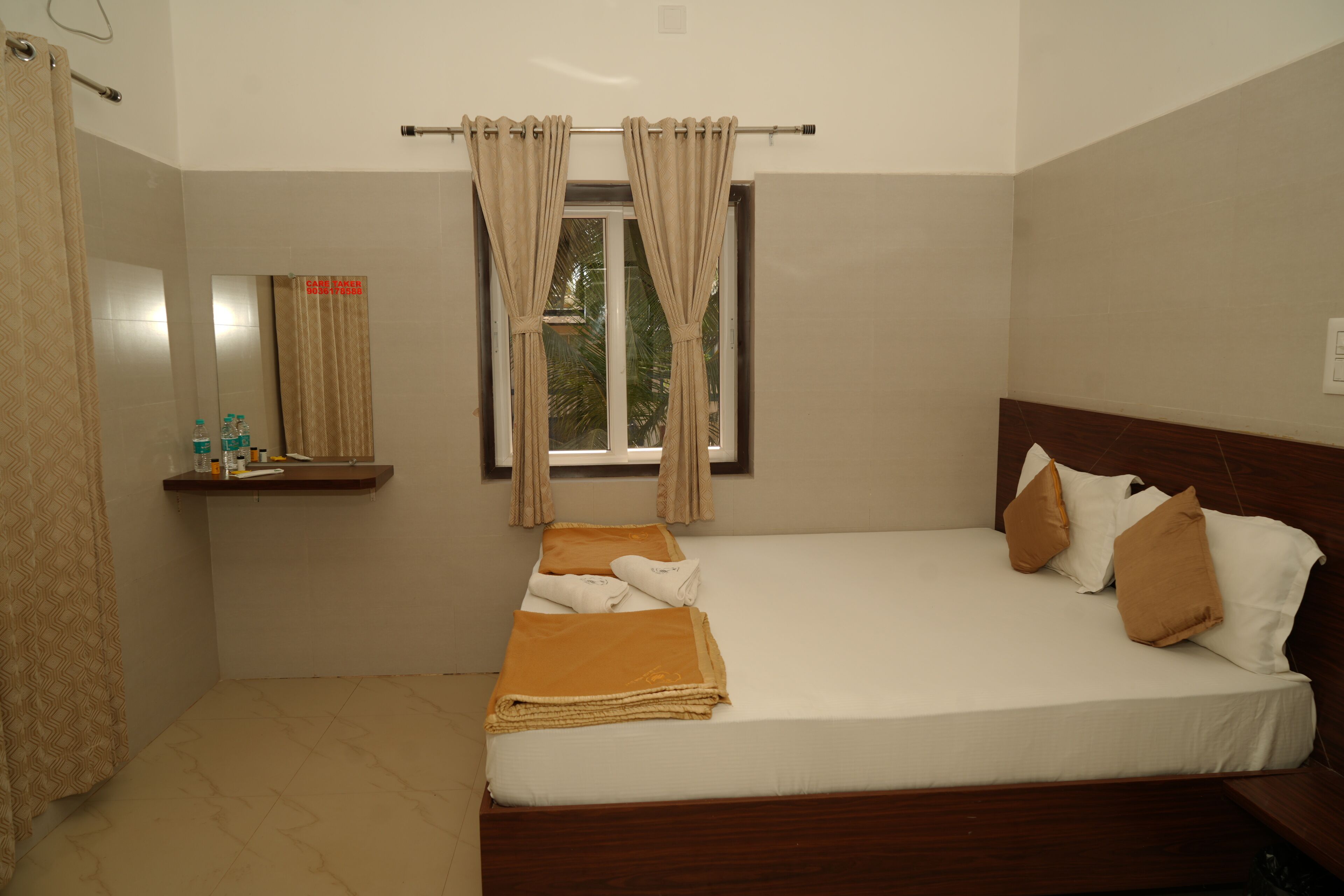 Habitación de hotel con cama, ventana con cortinas, estante pequeño con artículos de aseo y espejo.