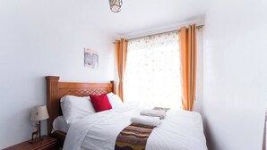 A neatly made bed with white linens and a red accent pillow, a wooden nightstand with a lamp, and a window with patterned curtains.