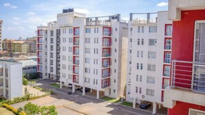 A multi-story residential building with balconies, a parking area, and a clear sky.