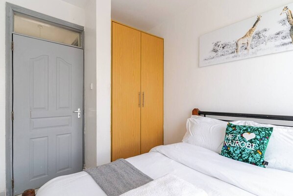 Un dormitorio con una puerta gris, un armario de madera, una cama con ropa blanca y una almohada con motivos de jirafa.
