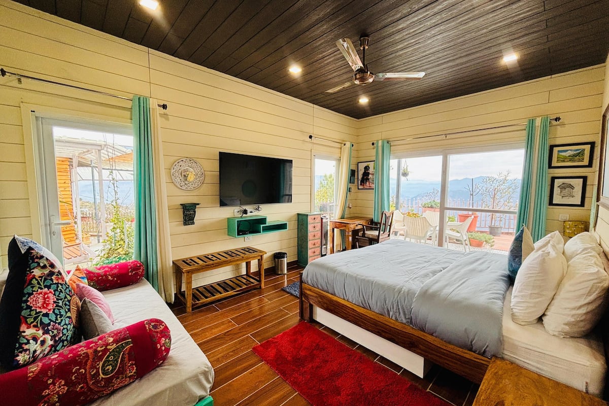 A bedroom with a wooden bed, a sofa with colorful cushions, a TV on the wall, and a view of the outdoors through large windows.