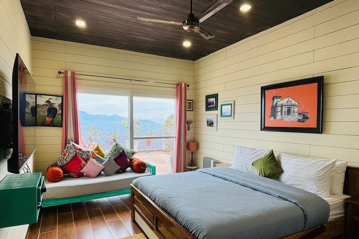 A bedroom with a bed, pillows, a window with a view, a ceiling fan, and wall-mounted pictures.