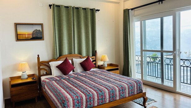 A bedroom with a wooden bed, green curtains, a nightstand with a lamp, a picture on the wall, and a balcony with a railing. - The Ridge House - A Villa Community (Tehri)