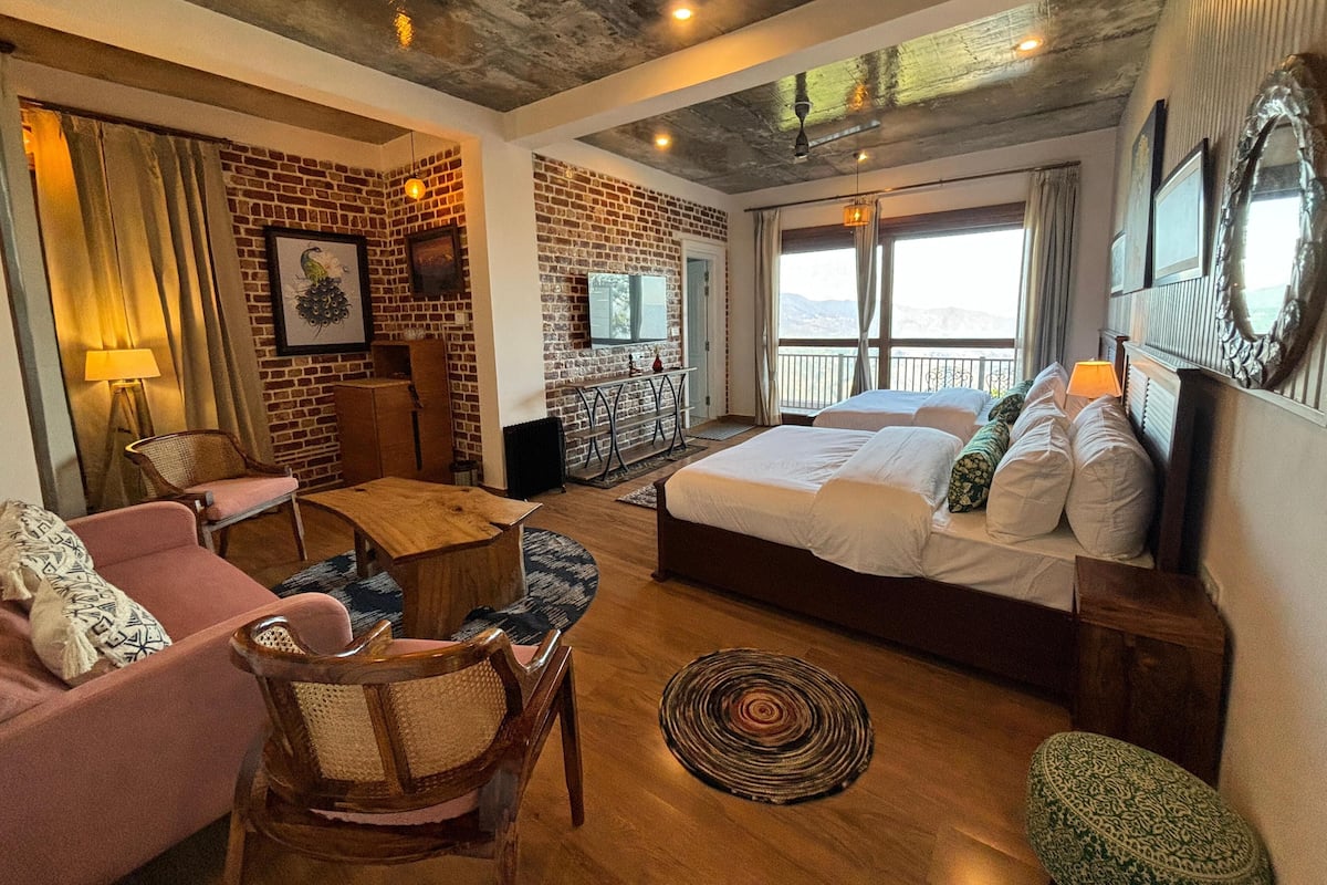 A hotel room with a brick wall, a bed with white linens, a wooden coffee table, a pink armchair, and a view of the outdoors.