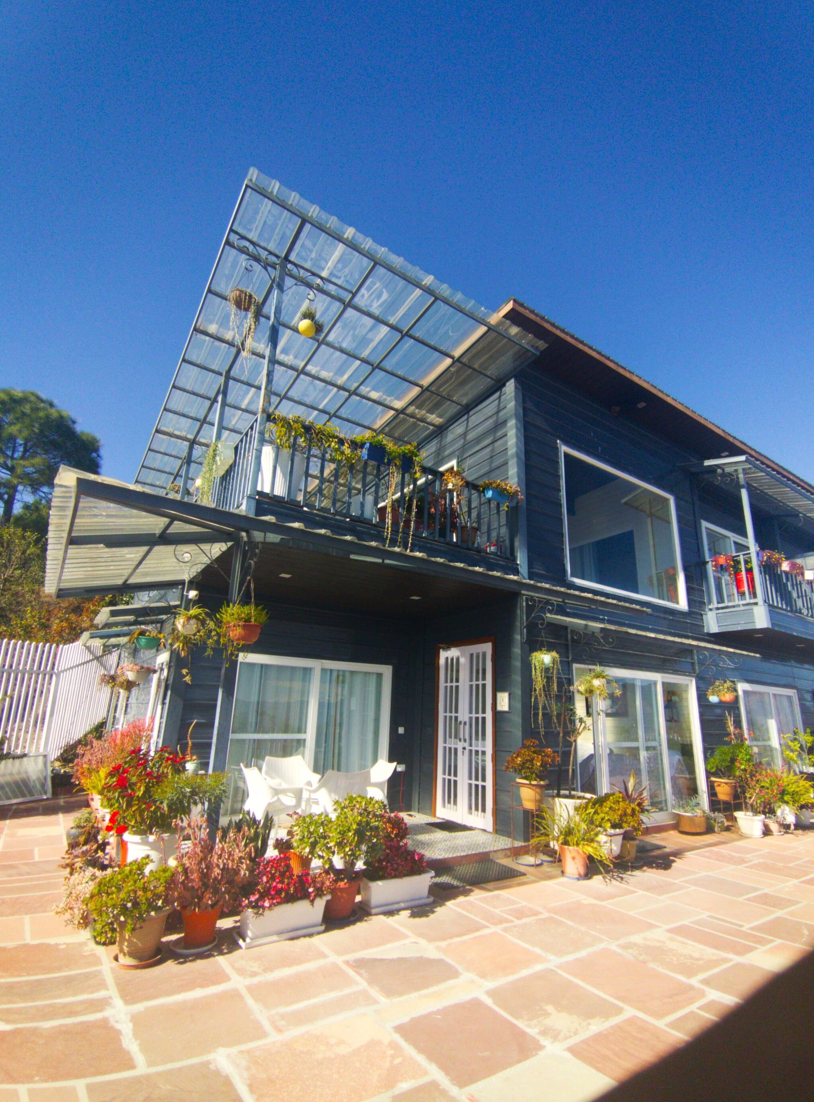 A two-story house with a large glass conservatory on the upper level, surrounded by potted plants and flowers.