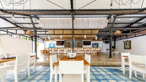 Un amplio comedor interior con sillas blancas y una mesa de madera, que cuenta con piso de baldosas y un techo alto con vigas a la vista.
