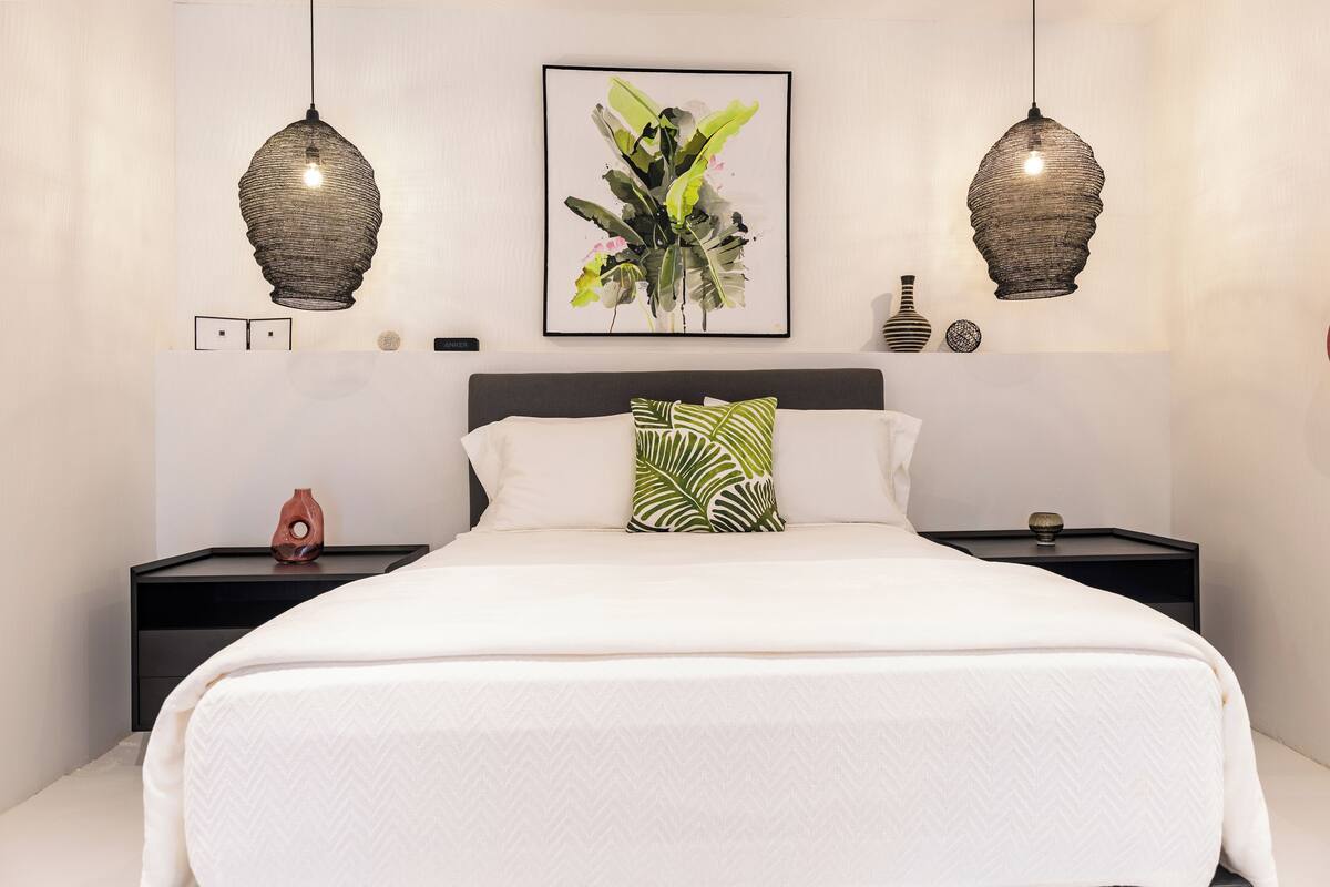 Un dormitorio con una cama grande, dos mesitas de noche, un velador, una planta enmarco y dos lámparas colgantes.
