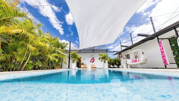 Una piscina con piso de baldosas, rodeada de palmeras y una pared blanca con una puerta y un banco.