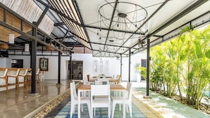 Amplio espacio interior con una larga mesa de comedor, sillas blancas y suelo de baldosas. Hay plantas a la derecha y un techo con vigas a la vista y luminarias.