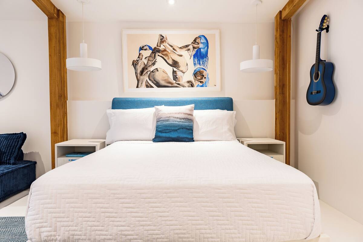 Un dormitorio con una cama grande, cabecera azul, dos mesitas de noche, una guitarra en la pared y un cuadro sobre la cama.