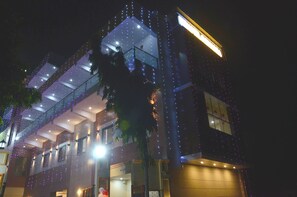 A multi-story building with balconies and a glass facade at night, illuminated by string lights. - Hotel Subhadra (Kannauj)