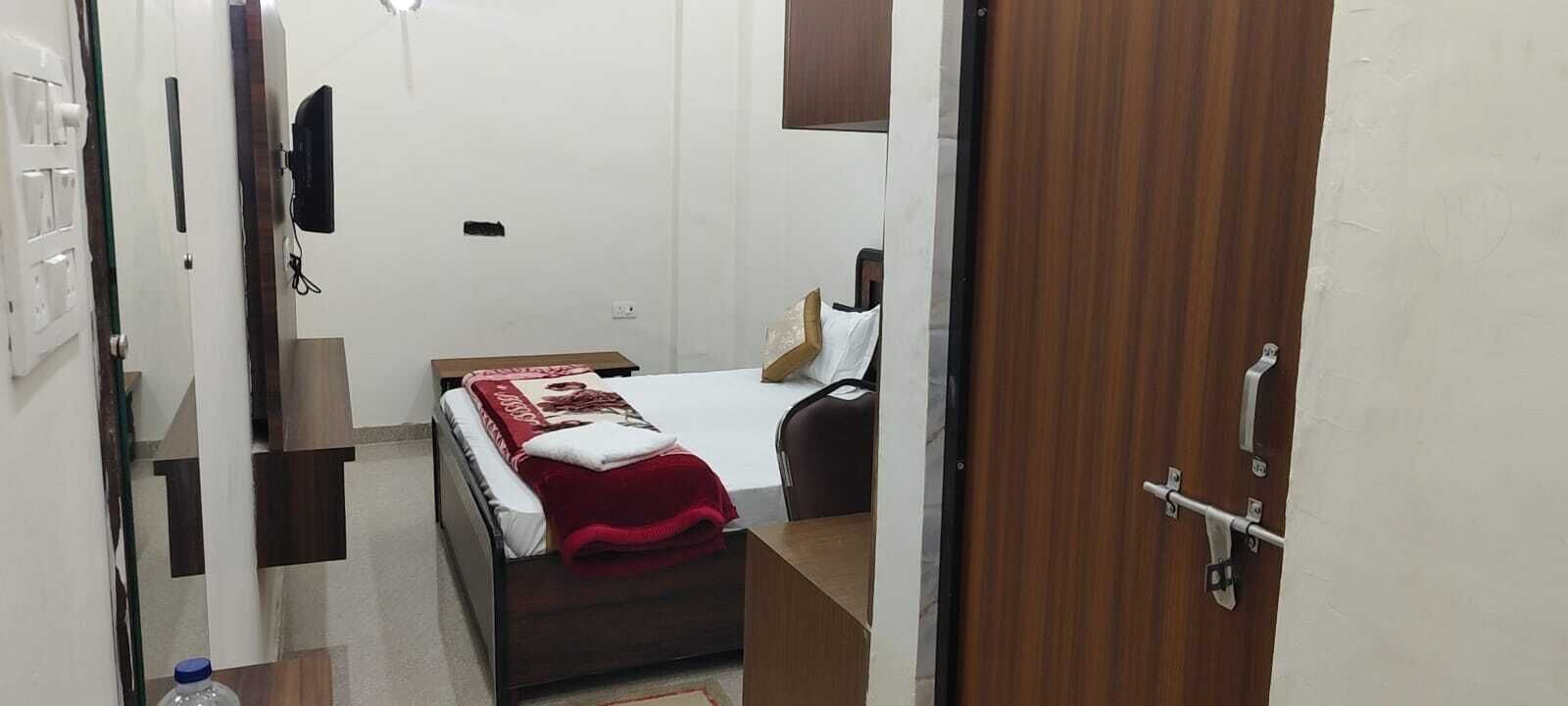 A small, compact room with a bed, a desk, and a chair. There is a television mounted on the wall and a door with a handle.