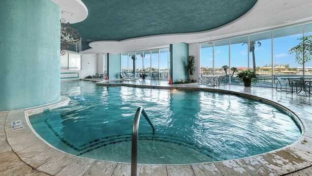 An indoor swimming pool with clear blue water, surrounded by large windows offering views of palm trees and buildings.