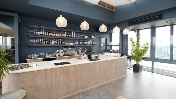 A modern bar area with a wooden counter, shelves of bottles, and hanging pendant lights.