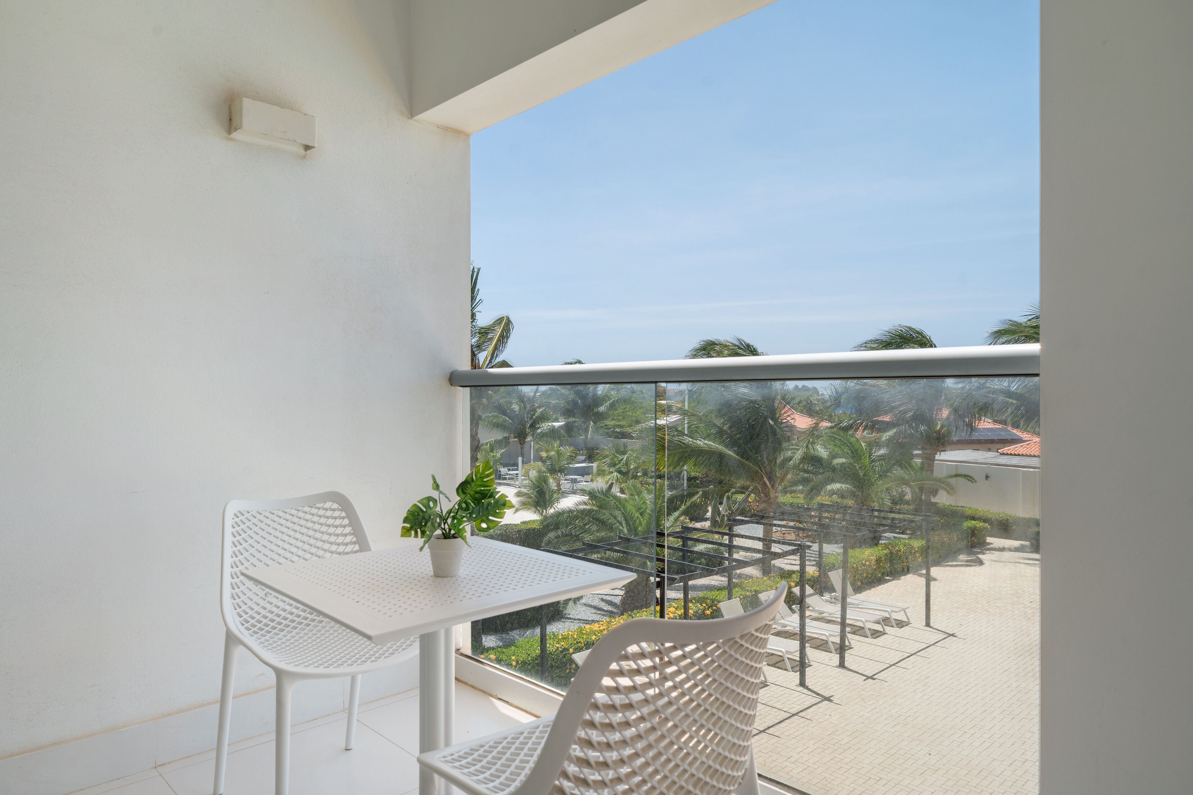 A balcony with a white table and chairs, a potted plant, and a view of a landscaped area with palm trees and a building in the background.