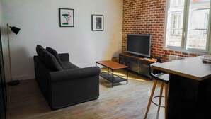 Una sala de estar con un sofá negro, una mesita de centro pequeña, un televisor sobre una pared de ladrillo y una ventana con persianas.