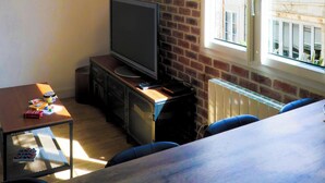 Una habitación con pared de ladrillo, un televisor sobre un mueble bajo, una mesa con objetos y una ventana con vistas a edificios.