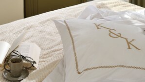 Ein Hotelzimmer mit Balkon, einem Bett mit weißer Bettdecke, einem Kopfkissen mit Initialen, einer Tasse und einem offenen Buch.