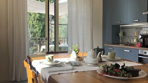 A dining area with a wooden table set for two, a kitchen with blue cabinets, and a view of greenery outside. - Elegant apartment, villa surrounded by greenery a stone's throw from the lake and the village (Garda)