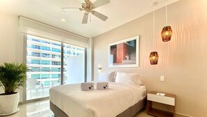 A modern hotel room with a large bed, a ceiling fan, a potted plant, a view of the city, and a small bedside table. - w Impeccable 2BR w Charming Balcony in CTG (Cartagena)