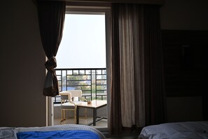A hotel room with a bed, a small table, and a chair. There is a balcony with a view of buildings and a railing.