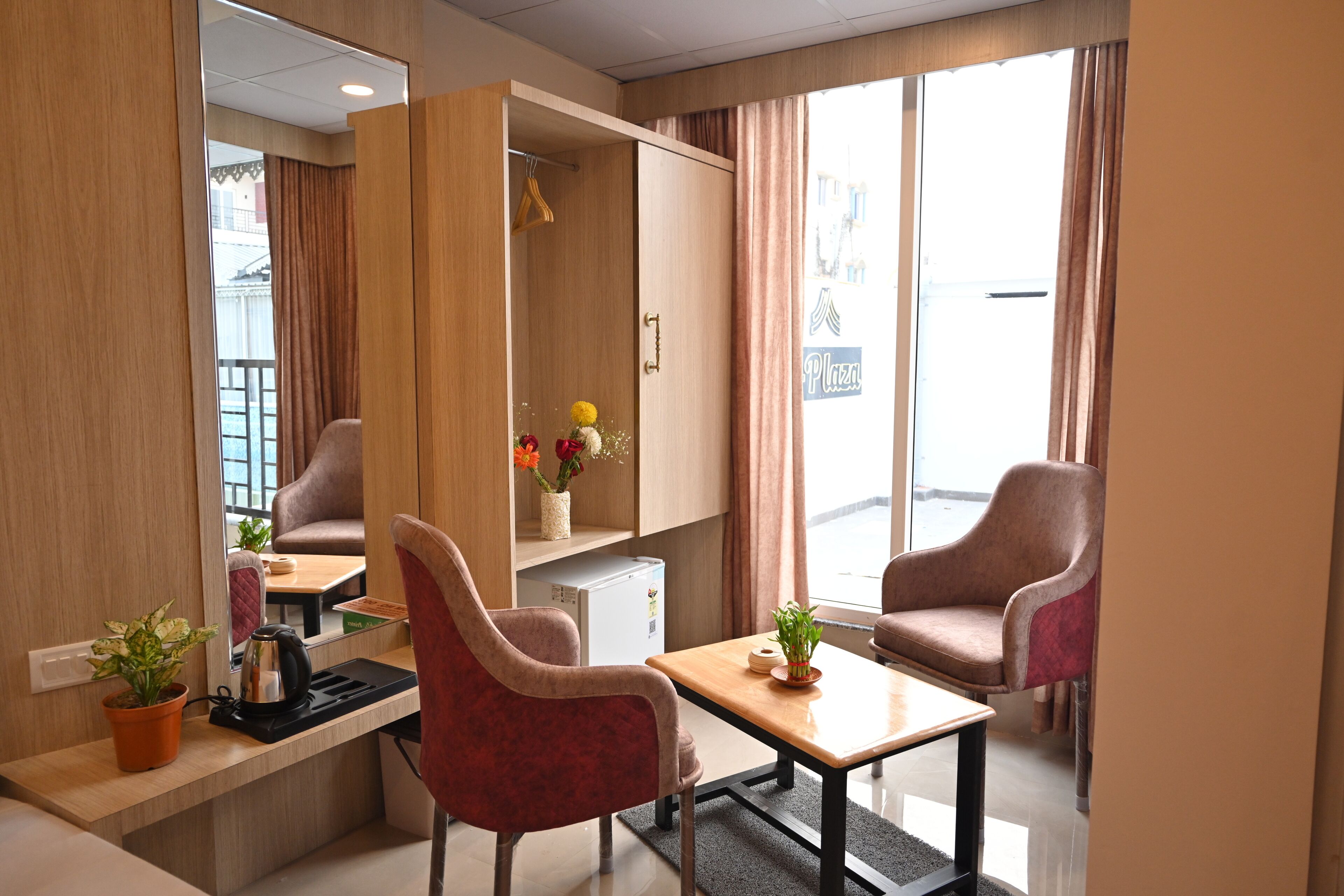A hotel room with a wooden wardrobe, two armchairs, a small table, a coffee machine, and a potted plant.