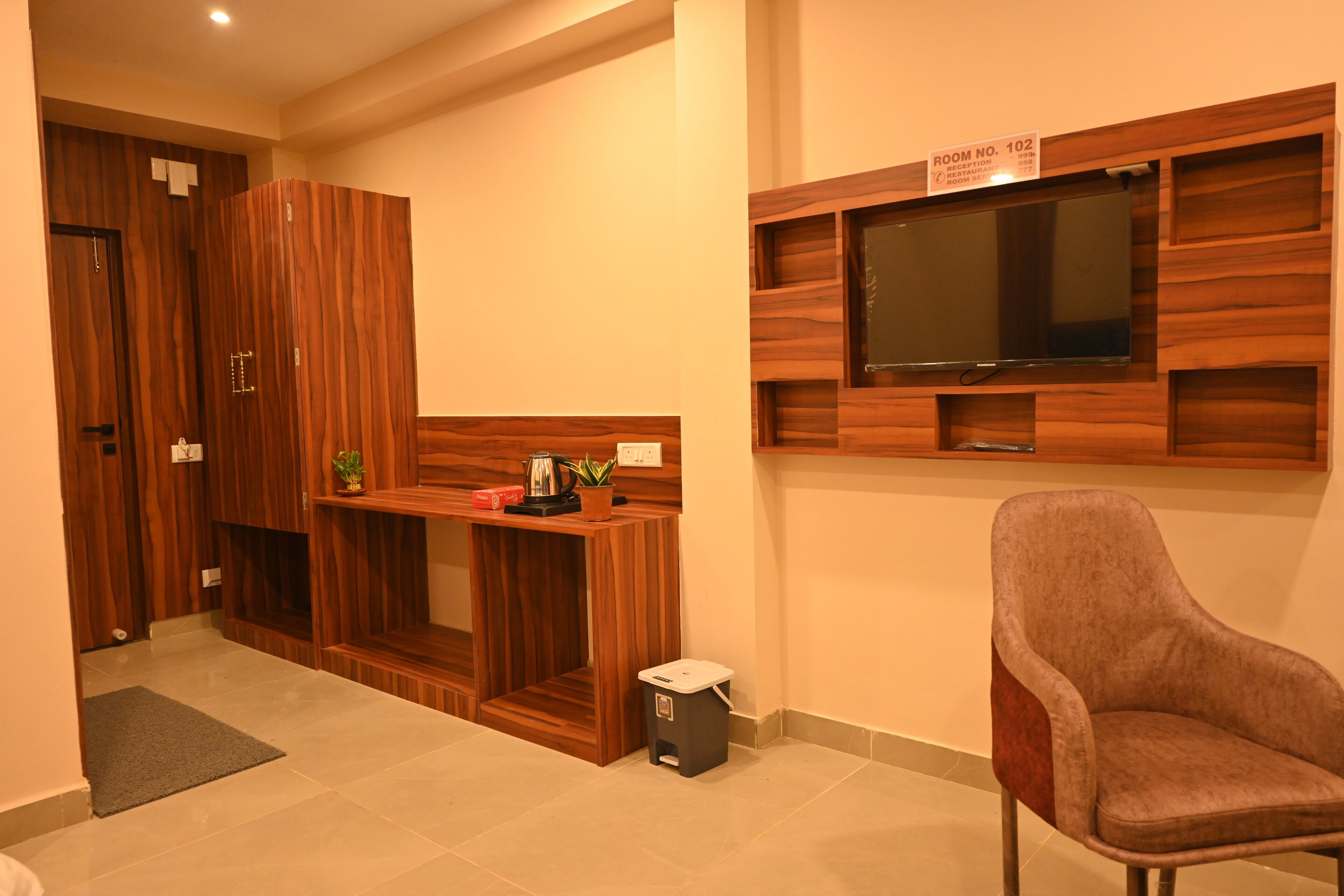 A room with a wooden wardrobe, a small table with a plant, a trash bin, and a TV mounted on the wall.