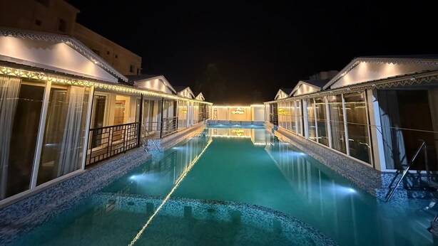 Vista nocturna de un hotel con piscina central, habitaciones comunicadas y una fachada bien iluminada.